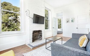 Ein modernes Wohnzimmer mit Kamin aus Stein, einem flachbildfernseher an der Wand, einem grauen Sofa mit Kissen und Holzboden.