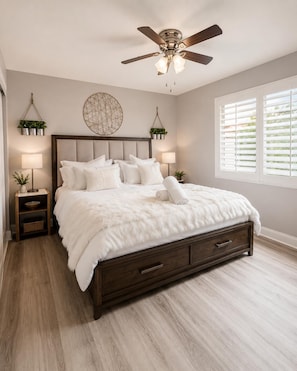 Une chambre à coucher comprenant un lit, un ventilateur de plafond, des tables de chevet et une fenêtre avec des stores.