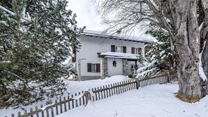 A snow-covered house with a stone foundation, wooden shutters, and a wooden fence. - Chesa Quadrellas / EG by Interhome (Samedan)