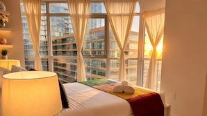 A hotel room with a large window, a bed with white linens, a red throw blanket, and a lamp on a bedside table.