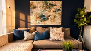 A living room with a sofa, cushions, a rug, a plant, and a large abstract painting on the wall.