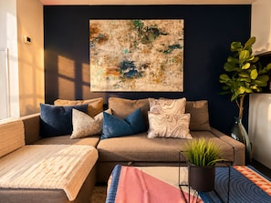 Ein Wohnzimmer mit einer Couch, Kissen, einem Teppich, einer Pflanze und einem großen abstrakten Gemälde an der Wand.