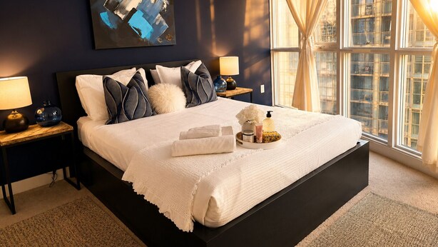 A modern hotel room with a large bed, two bedside tables, a nightstand, a window with curtains, and a cityscape view.