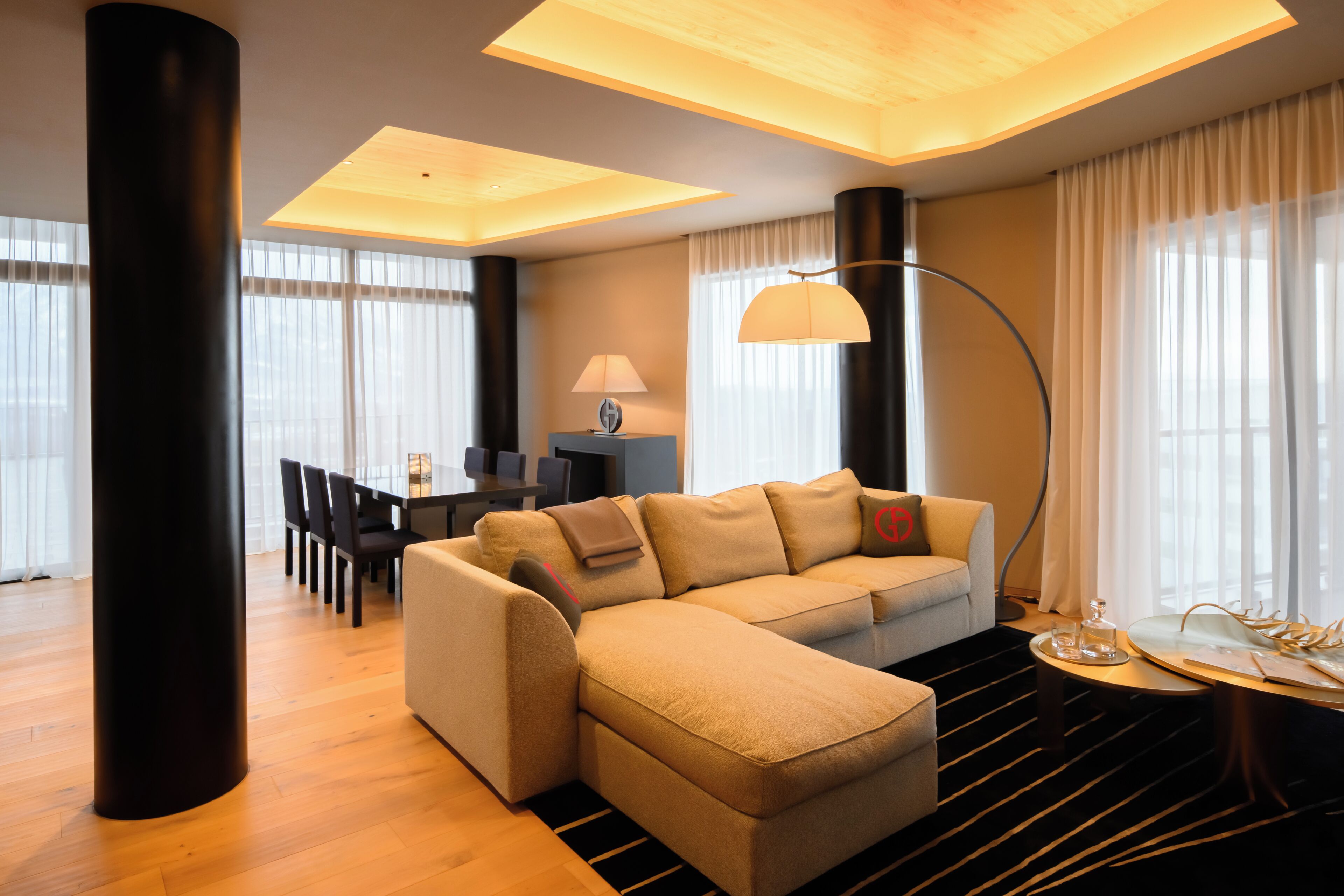A modern living room with a sectional sofa, a dining table with chairs, a lamp, and a view of the outdoors.