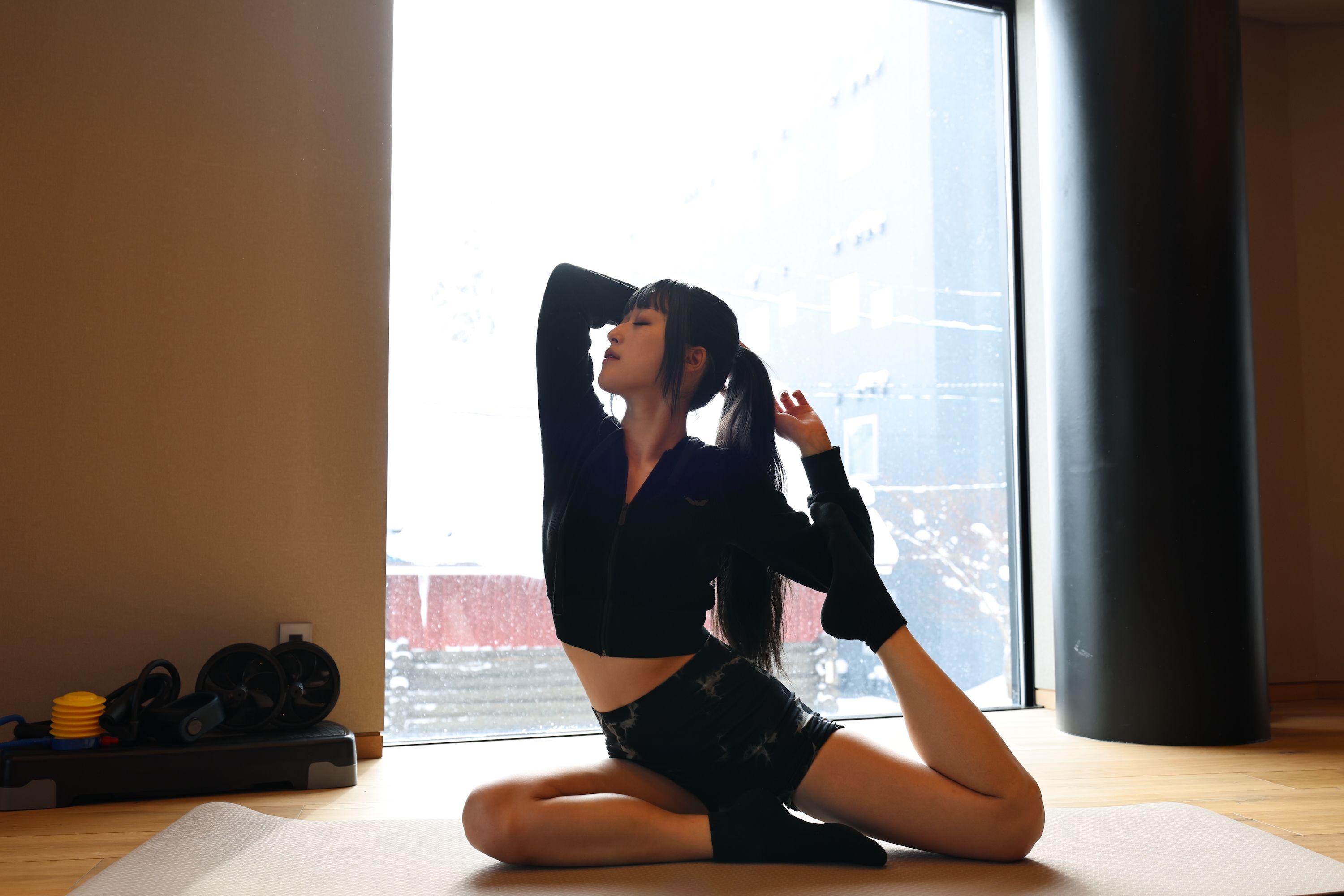 A person performing a yoga pose on a mat in a room with a large window, a punching bag, and a set of dumbbells.