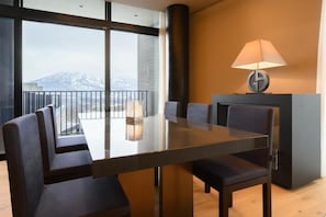 A modern dining area with a large table and chairs, a fireplace, and a view of mountains through large windows. - HOTELA Niseko Towers  (Kutchan)