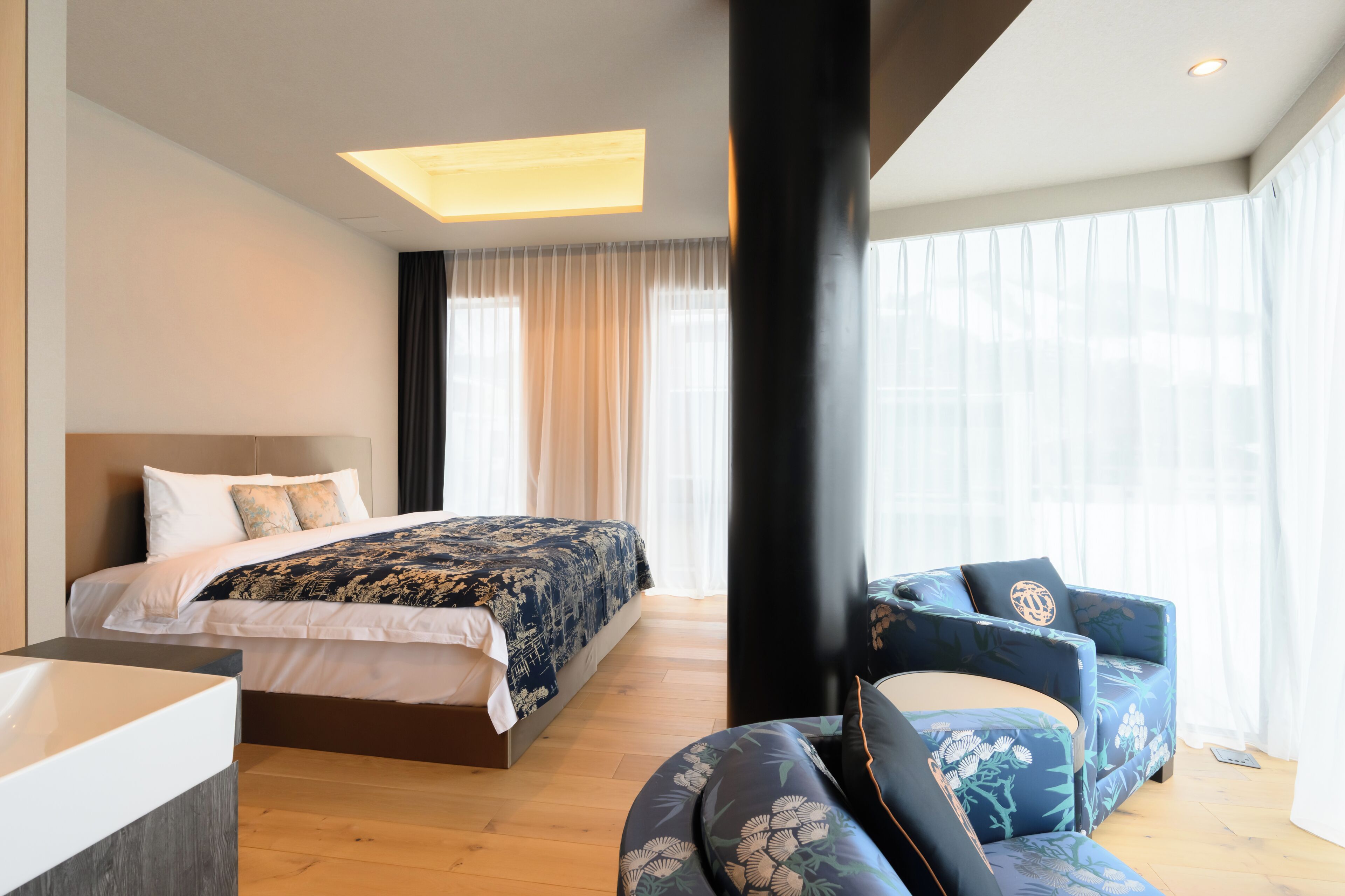 A modern hotel room with a large bed, a sitting area with a chair, and a large window with curtains.