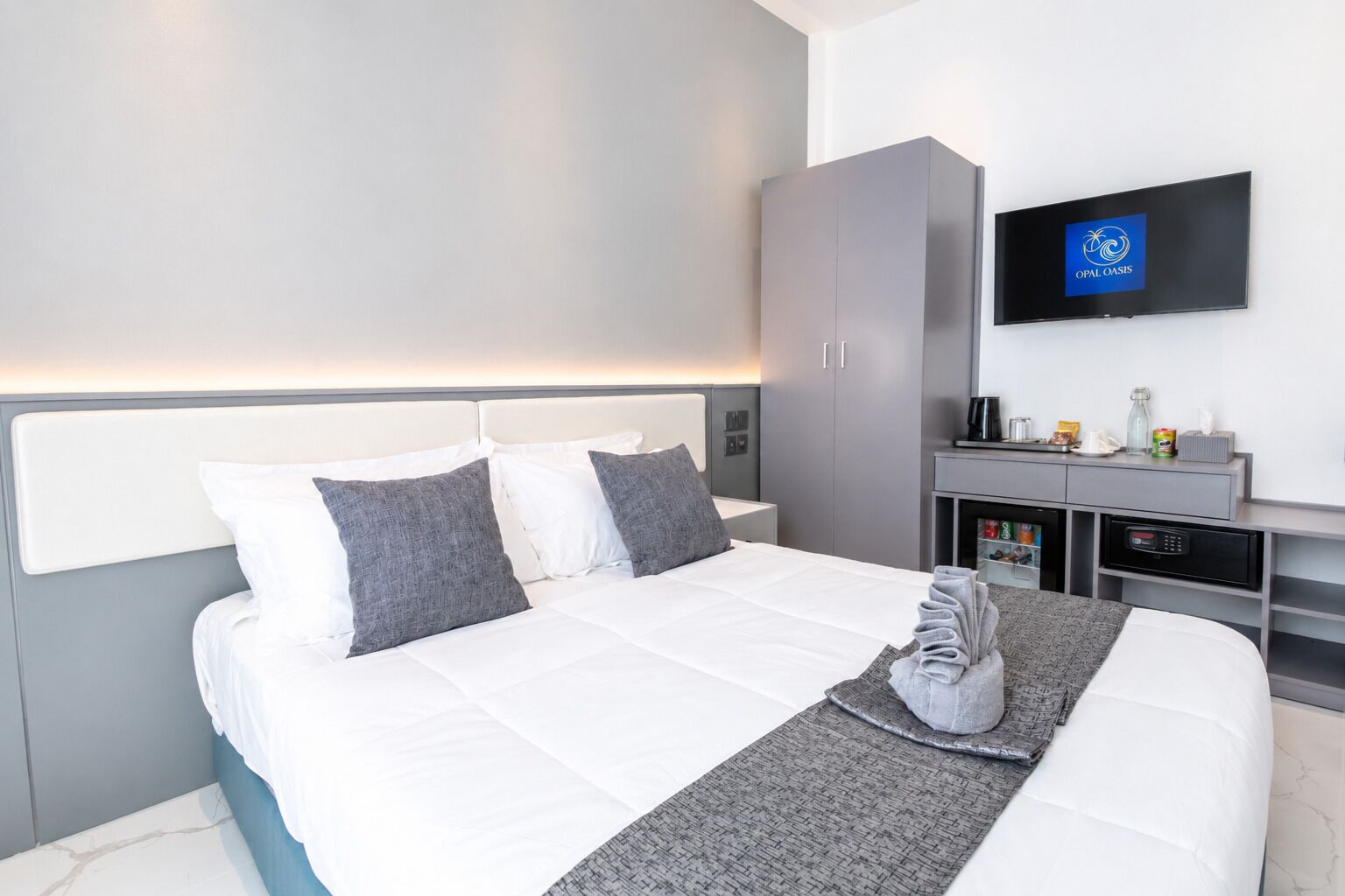 Una habitación de hotel moderna con una cama grande, ropa de cama gris y blanca, un televisor de pantalla plana, un armario y una pequeña mesa de centro con artículos de cortesía.