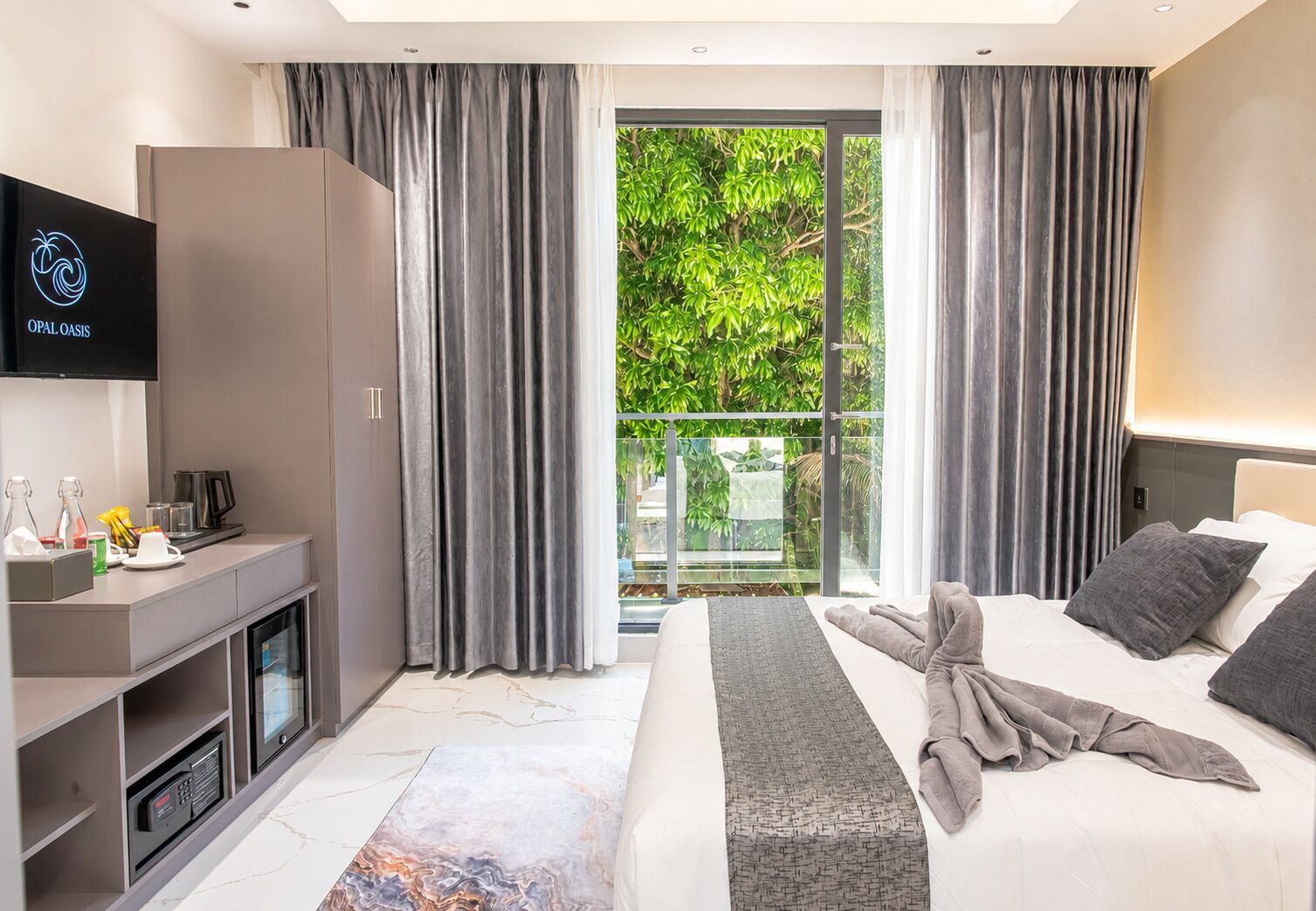 A modern hotel room with a bed, a TV, a minibar, and a balcony with a view of greenery.