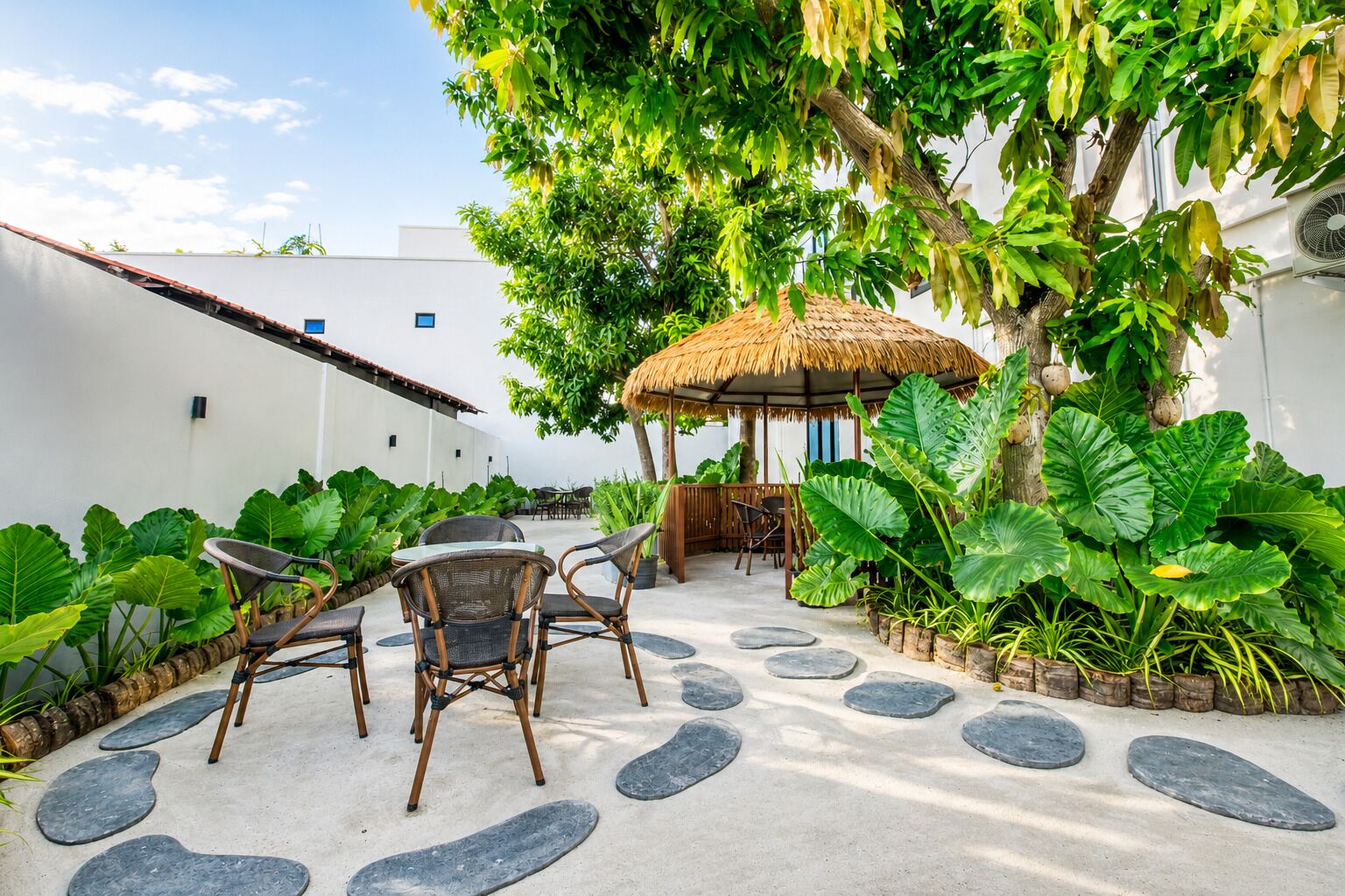 An outdoor seating area with a thatched-roof gazebo, surrounded by tropical plants and a stone pathway.