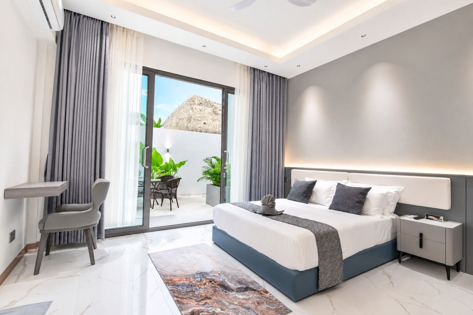 A modern hotel room with a bed, bedside tables, a desk, and a balcony with plants.