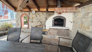 Eine Terrasse im Freien mit Steinmauern, einem Backofen aus Ziegeln, Metallstühlen und einem Holztisch.