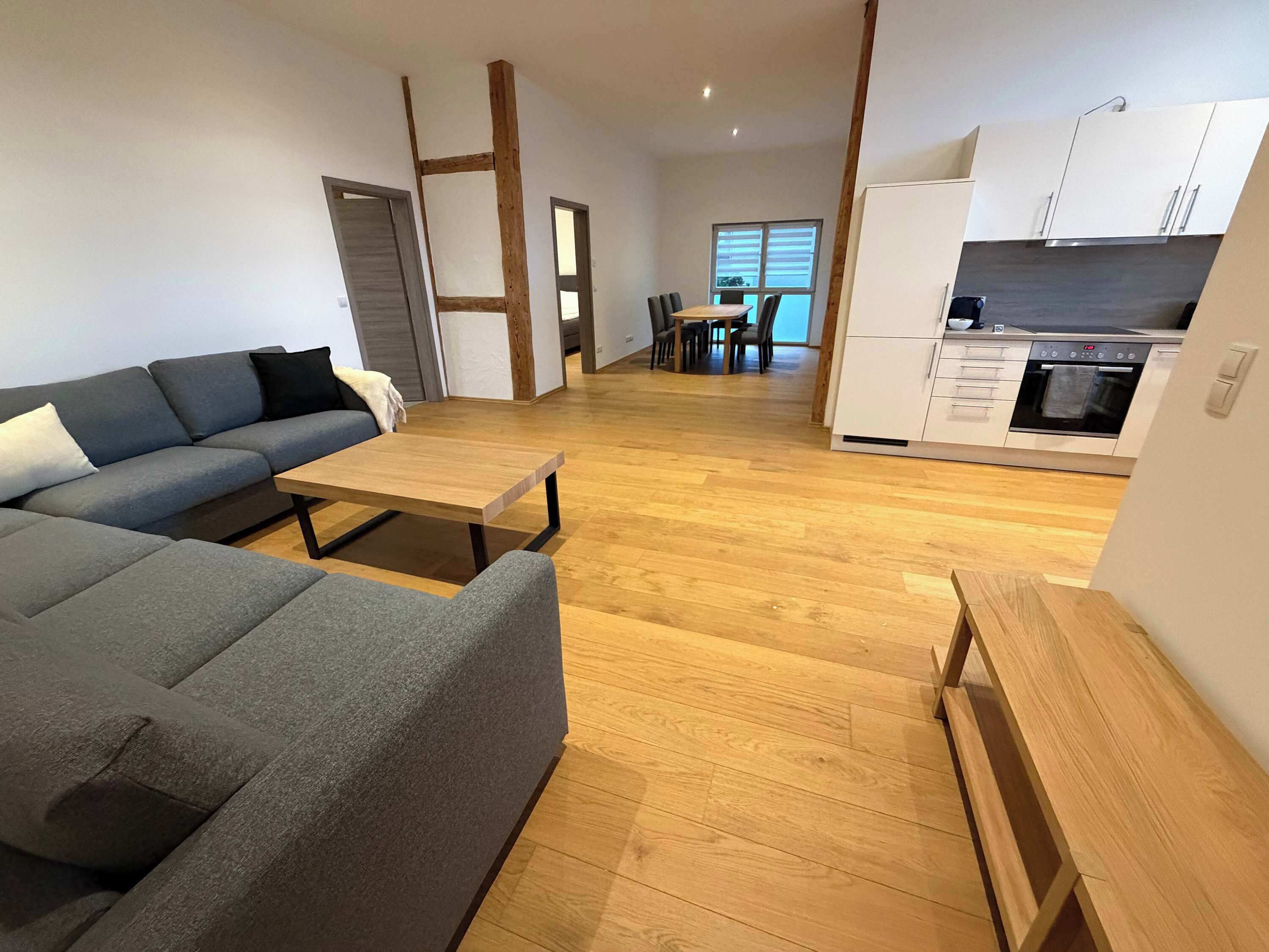 Ein modernes Wohnzimmer mit grauem Sofa, einem Holztisch und einer Küchenzeile mit weißen Schränken und einem Backofen.