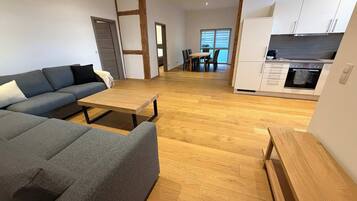 Ein modernes Wohnzimmer mit grauem Sofa, einem Holztisch und einer Küchenzeile mit weißen Schränken und einem Backofen.