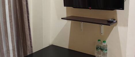 Una habitación pequeña con una mesa negra, una silla, un televisor montado y un estante con dos botellas de agua.