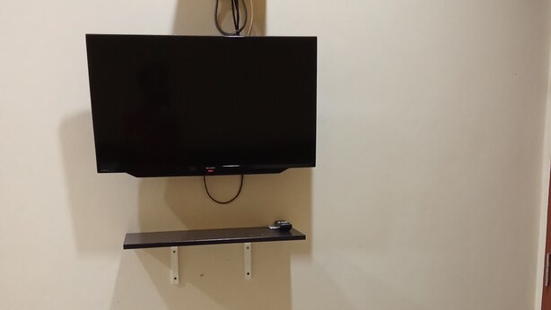 A wall-mounted television with a stand below it, a power outlet above, and a door to the right. - Tanjong Inn by StayZy (Kota Bharu)