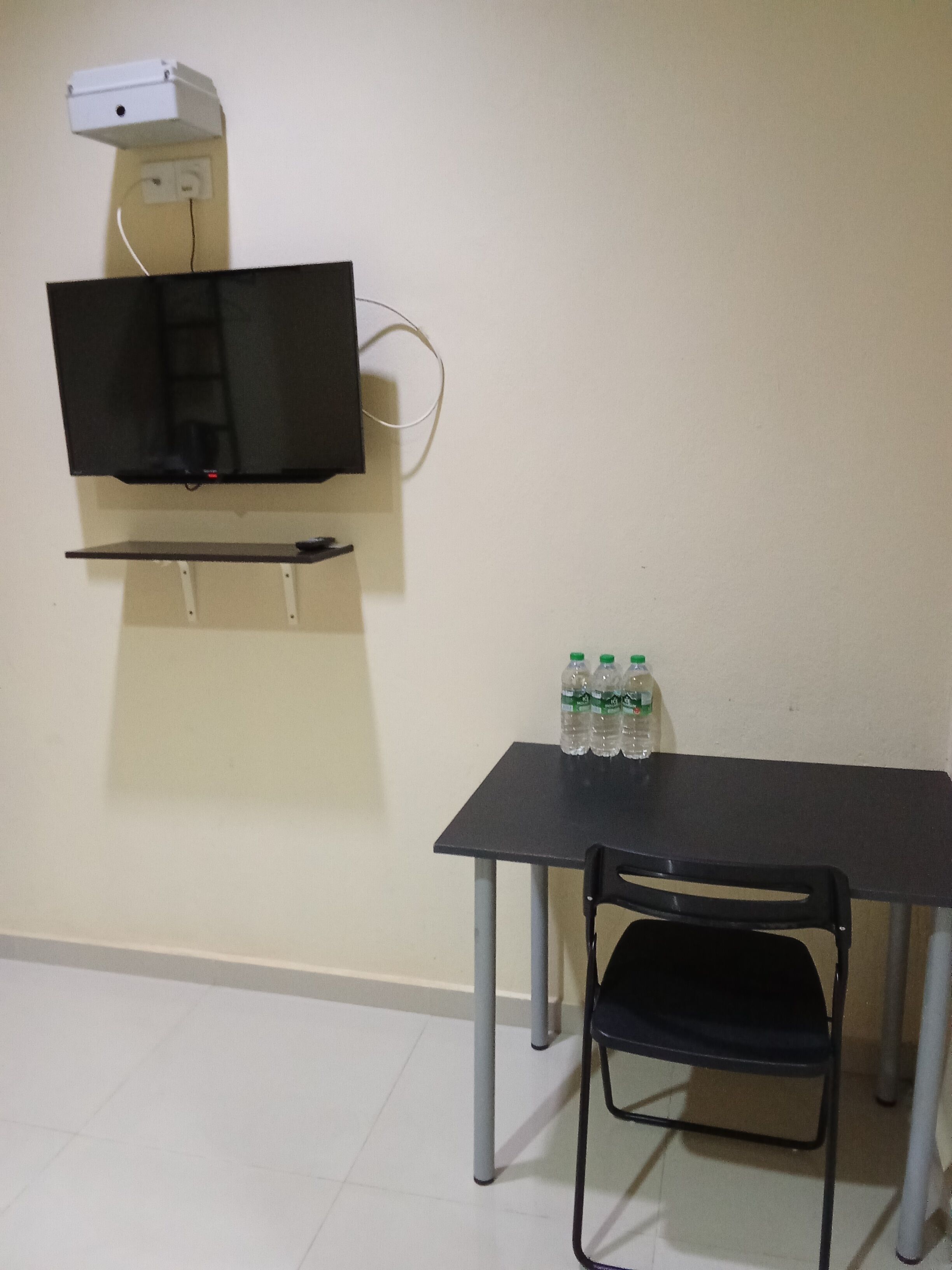 A room with a flat-screen TV mounted on the wall, a small table with a chair, and a shelf with bottled water.
