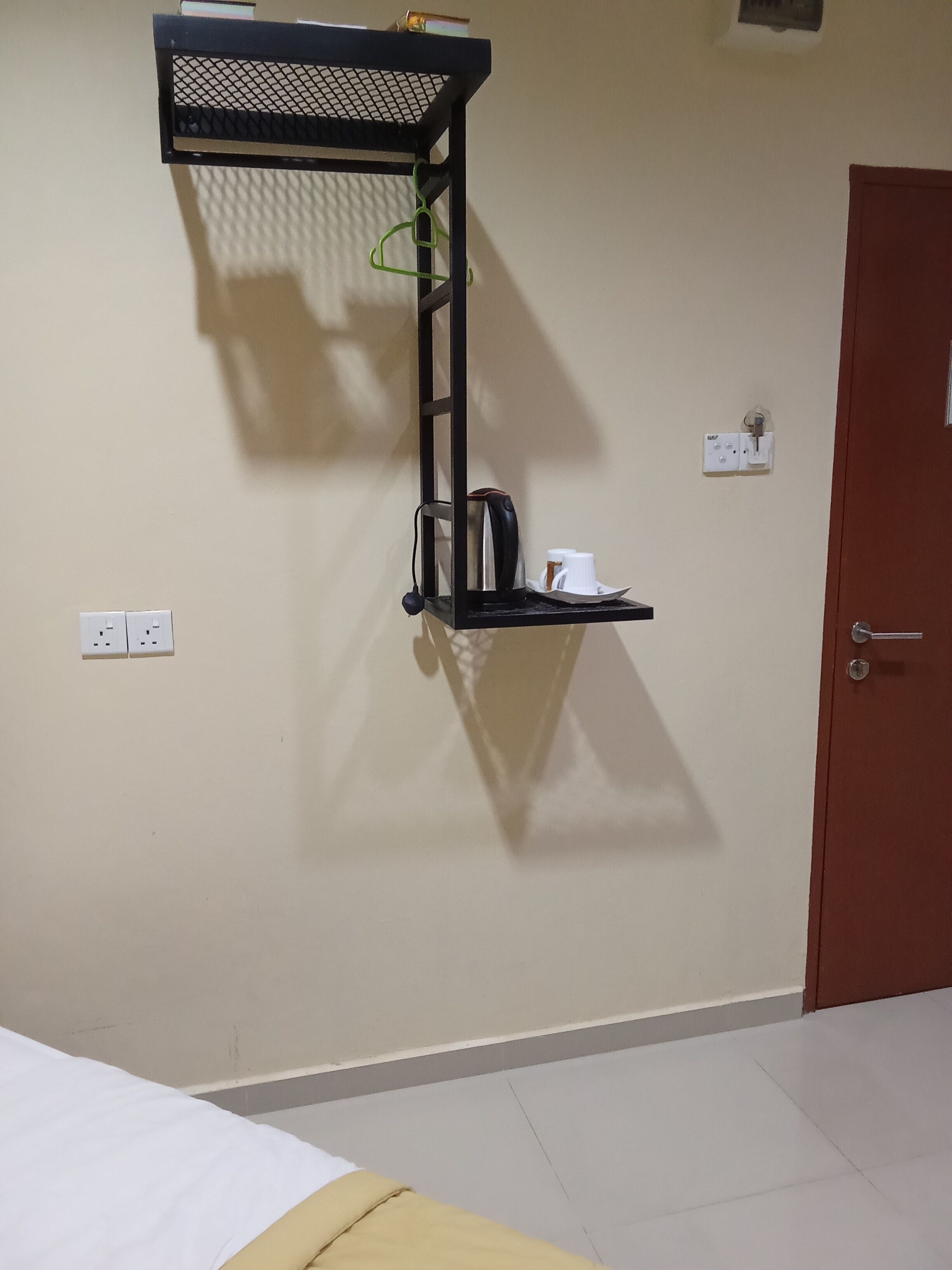 A room with a bed, a wall-mounted shelf with a kettle and cups, and a door.