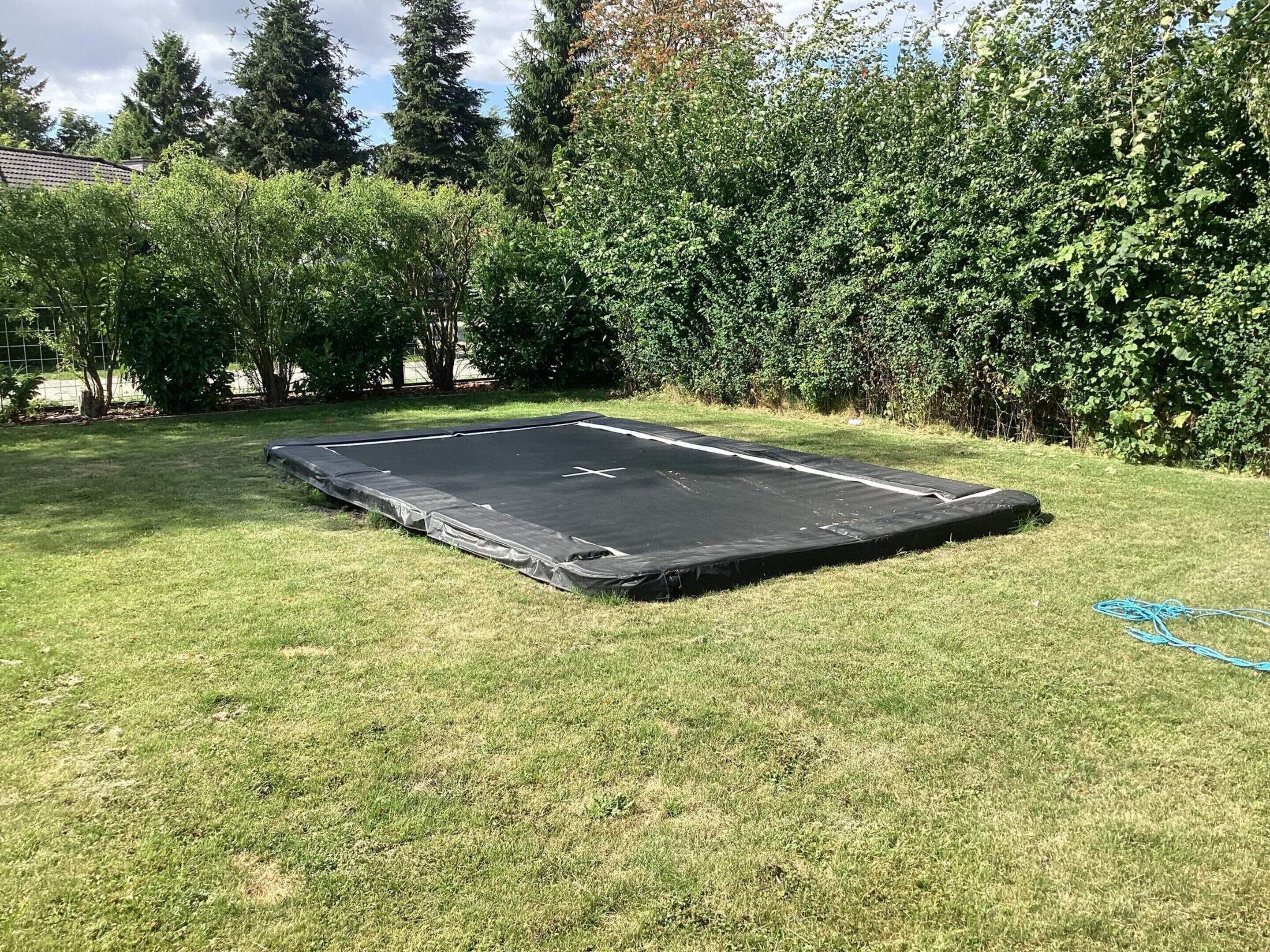 Un trampoline se trouve dans une cour verdoyante, avec des arbres et une haie en arrière-plan.