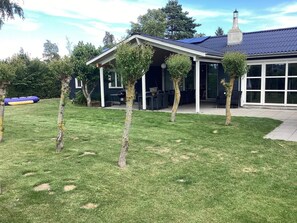 Ein Haus mit überdachter Terrasse, Schornstein und einem Rasen mit jungen Bäumen.