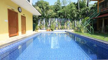 A swimming pool with a tiled edge, surrounded by a building with a reception and kitchen area, and a lush green backdrop.
