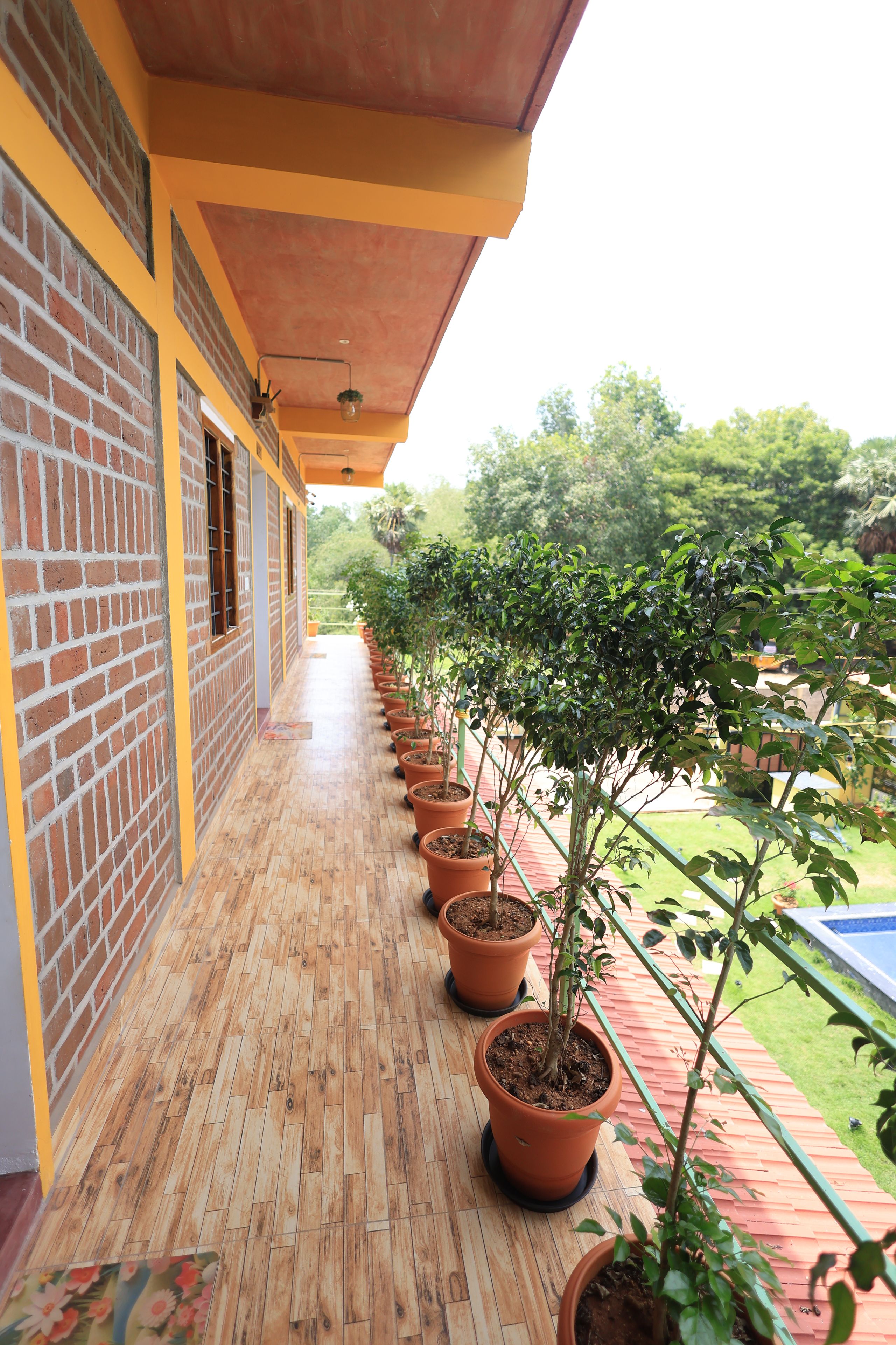 A covered balcony with a row of potted plants, a brick wall, and a wooden floor.