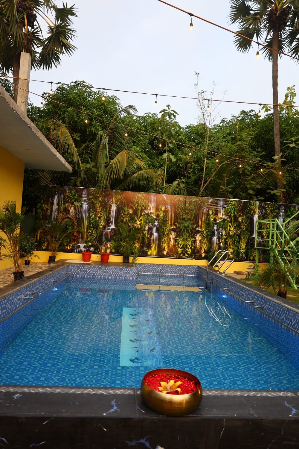 A swimming pool with a waterfall feature, surrounded by lush greenery and potted plants.