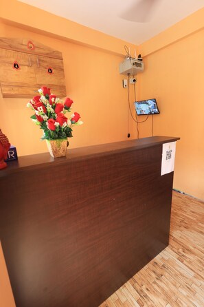 Un mostrador de recepción con detalles de madera, un arreglo floral rojo en maceta y una cámara de seguridad montada.