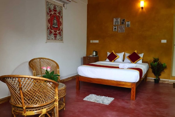A hotel room with a bed, a chair, a small table, a wall tapestry, and a wall-mounted light.
