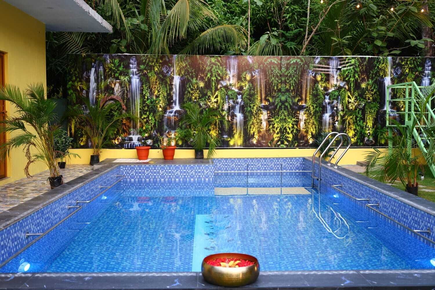 Une piscine dont les bords sont recouverts de carreaux, entourée de plantes en pot et bordée d’un mur orné d’une fresque représentant une cascade.
