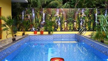 Une piscine dont les bords sont recouverts de carreaux, entourée de plantes en pot et bordée d’un mur orné d’une fresque représentant une cascade.