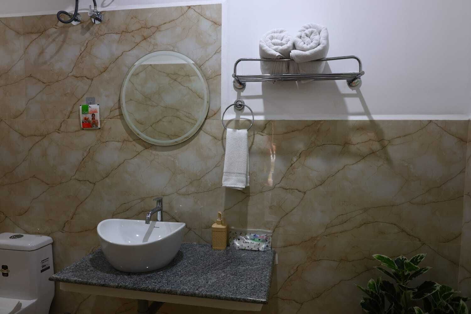 Un baño con pared de mármol, un espejo redondo, un lavabo con grifo, un perchero con toallas y un dispensador de jabón.