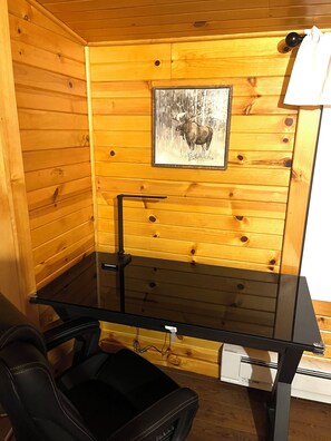 A wooden cabin interior with a framed moose painting on the wall, a black chair, and a small table.