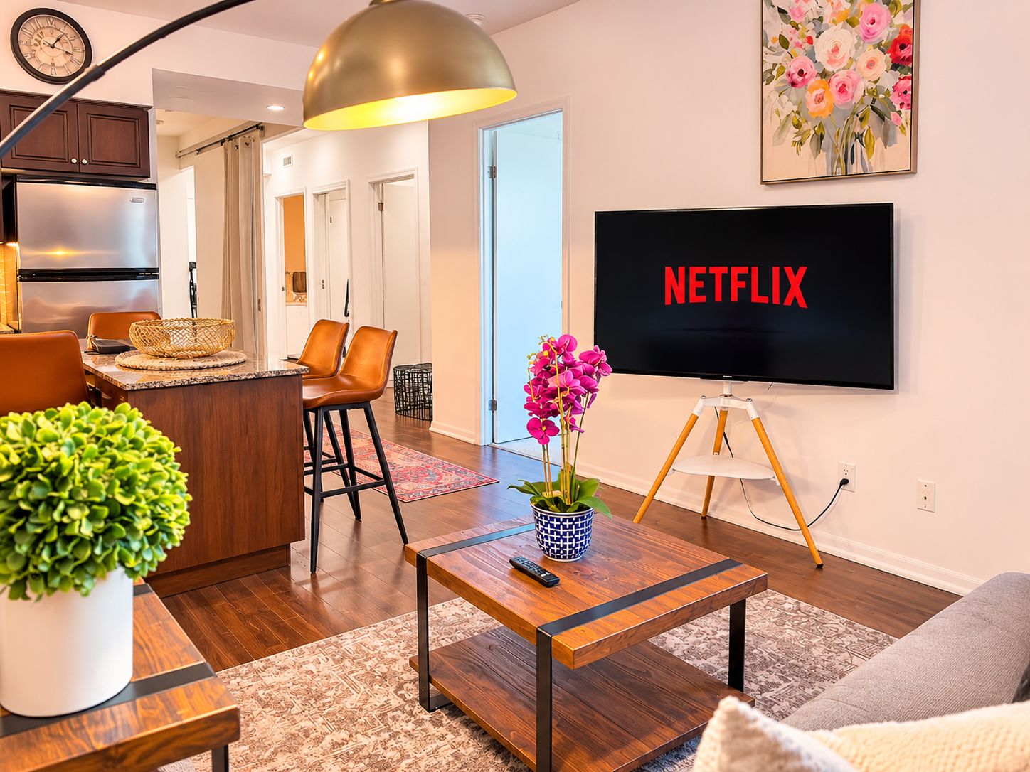 A modern living room with a flat-screen TV displaying 'NETFLIX,' a wooden coffee table with a remote control, a potted plant, and a dining area with chairs and a basket.