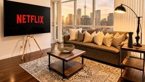 Een woonkamer met een grote tv waarop 'NETFLIX' staat, een bank met kussens, een salontafel, een lamp en uitzicht op een stadshemelbeeld.