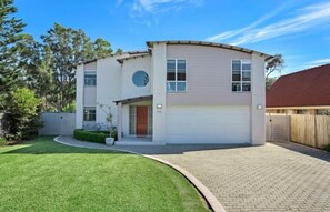 A two-story house with a garage, a circular window on the second floor, and a well-maintained lawn. - Ultimate Beach Escape: Family Haven, Firepit, near Café, Pets welcome, Sleeps 8 (Culburra Beach)
