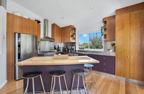 A modern kitchen with a central island, bar stools, wooden cabinets, stainless steel appliances, and a view through the window. - Ultimate Beach Escape: Family Haven, Firepit, near Café, Pets welcome, Sleeps 8 (Culburra Beach)