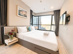 A modern hotel room with a large bed, a bedside table with a vase of flowers, a flat-screen TV mounted on the wall, and a window offering a city view. - Sea View 2 Bed Room Nice Location (Pattaya)