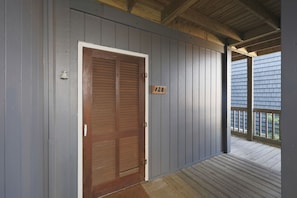 A wooden door with a number '128' on a gray wall, leading to a porch with a wooden railing. - Water's Edge | Coastal Condo with Ocean Views (North Topsail Beach)