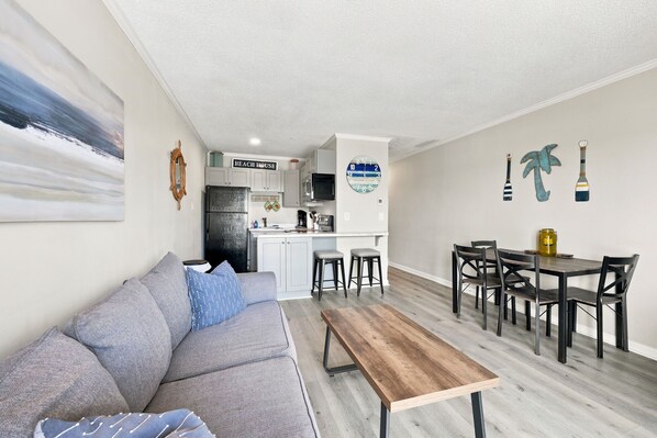 A modern living room with a sofa, coffee table, and a dining area with chairs and a table. - Water's Edge | Coastal Condo with Ocean Views (North Topsail Beach)