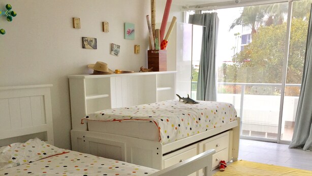 Un dormitorio con dos camas individuales, un armario blanco, una mesita con un florero y vistas al exterior a través de un ventanal.