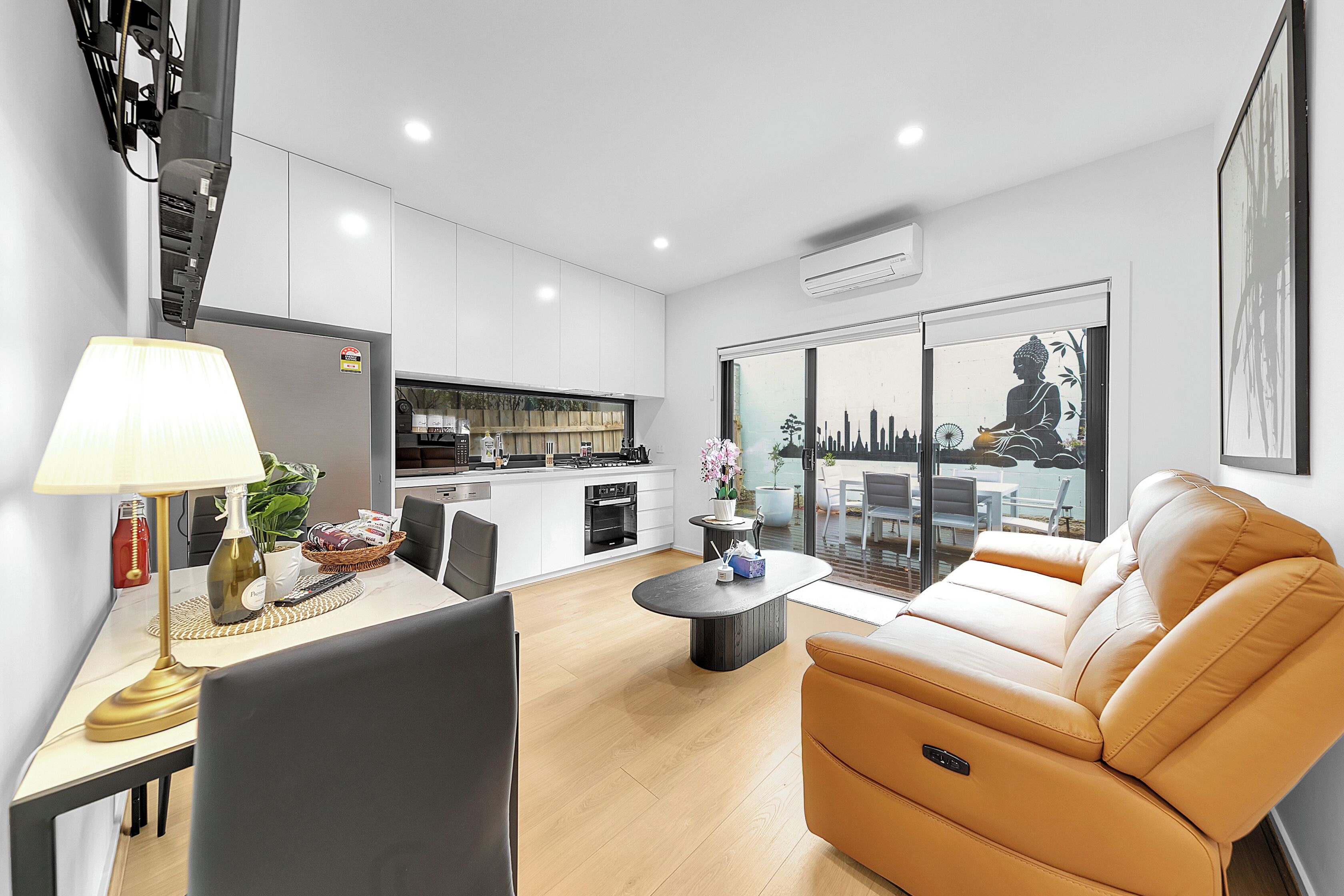 A modern living room with a sofa, a dining area with a table and chairs, a kitchen with white cabinets, and a large window with a cityscape view.