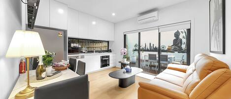A modern living room with a sofa, a dining area with a table and chairs, a kitchen with white cabinets, and a large window with a cityscape view.