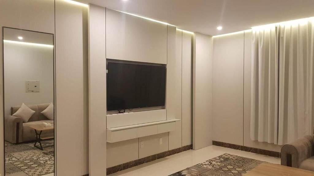 Een moderne woonkamer met een flatscreen televisie aan een witte muur, een bank en een kamerbreed tapijt met een patroon.