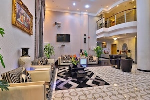Un hall de hotel con techo alto, una zona de estar central con sofás y sillones, piso estampado y una escalera que lleva a un nivel superior.