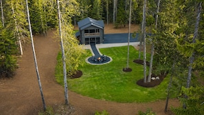 A lodge with a central fire feature surrounded by outdoor seating, set in a forested area with a paved driveway leading to the entrance.