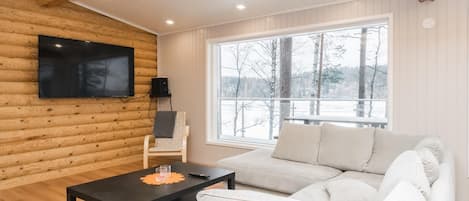 Una sala de estar moderna con una pared de troncos de madera, un televisor de pantalla plana, un sofá y un ventanal con vistas a árboles.