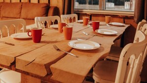 Mesa de madera dispuesta con platos, tazas y cubiertos, rodeada de sillas blancas.