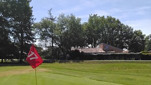 A golf course with a red flag marker on the green, surrounded by trees and a building in the background. - Maison avec piscine sur un golf (Fiac)