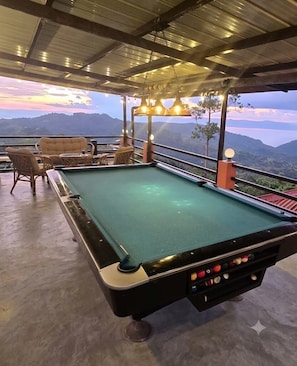 A pool table with surrounding chairs and tables, set against a scenic mountain backdrop.
